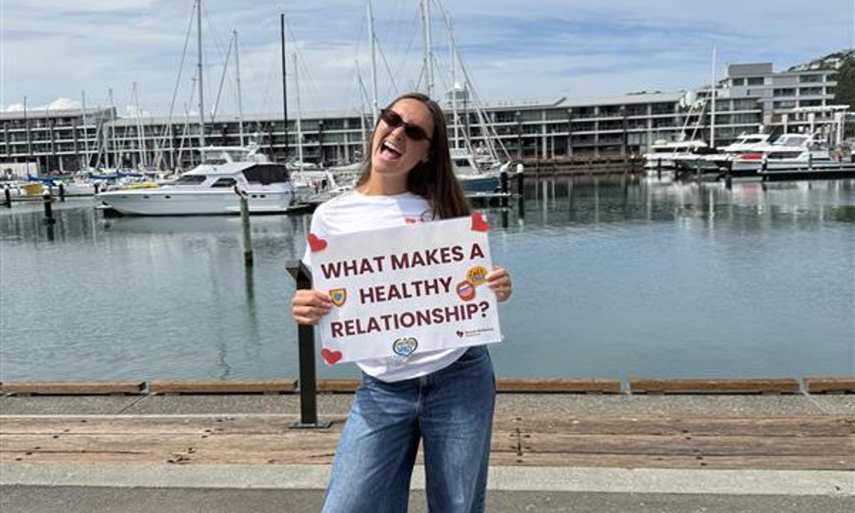 Photo of a woman standing by Wellington waterfront with a sign saying What makes a healthy relationship?